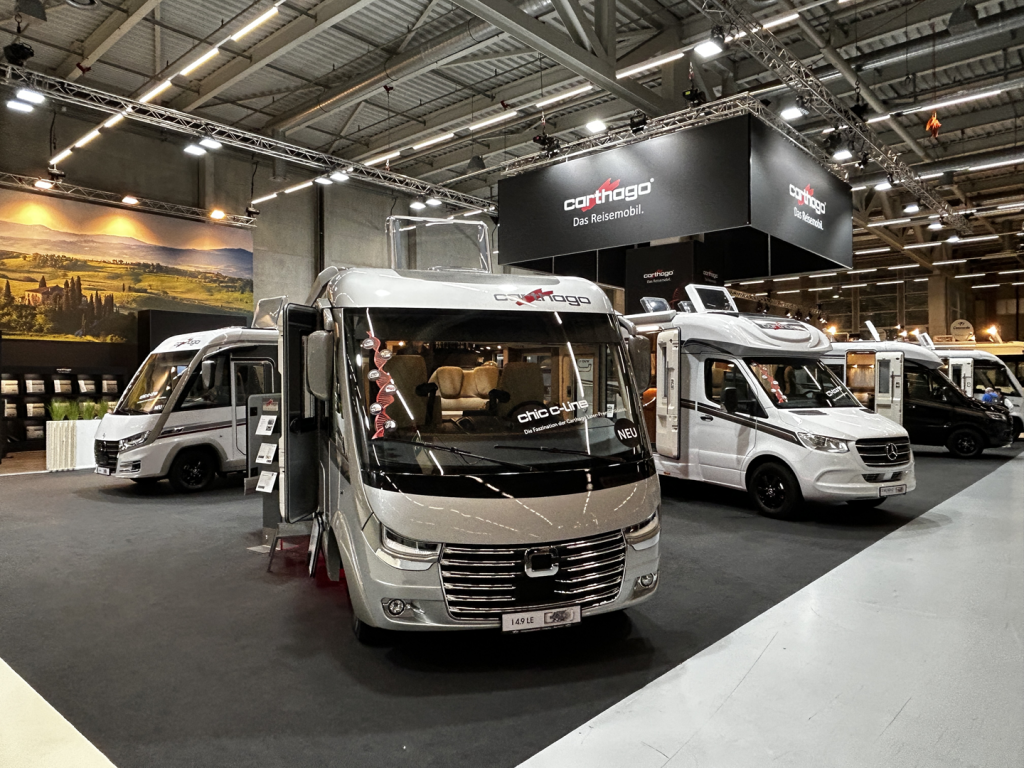 An einem überdachten Messestand mit Carthago-Branding sind drei Luxus-Wohnmobile ausgestellt. Der Bereich ist gut beleuchtet, und an der Rückwand ziert ein großes, malerisches Wandbild die Wand.