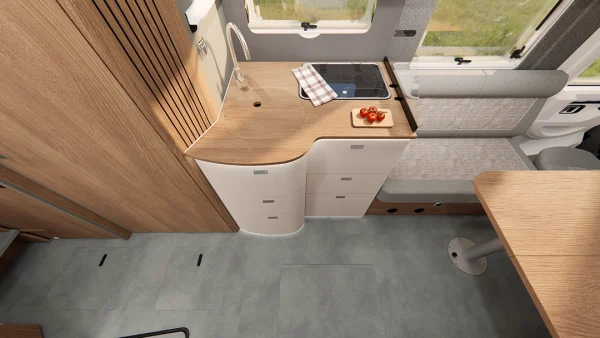Moderne Wohnmobilküche mit hellen Holzschränken, Spüle, Herd, kariertem Handtuch und einer kleinen Schale mit Äpfeln. Eine gepolsterte Sitzbank und ein Teil eines Tisches sind zu sehen. Die Fenster sorgen für Tageslicht und unterstreichen das durchdachte Design, das für einen Carthago C2-Tourer typisch ist.