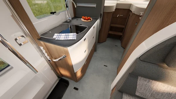 Kompakte Wohnmobilküche im Carthago C2-Tourer mit schwarzem Kochfeld, Spüle, Handtuch und einem Teller mit Tomaten auf der Arbeitsplatte. In dem gemütlichen Raum sind Holzschränke, ein grauer Boden und eine Treppe zu sehen, die nach oben führt.