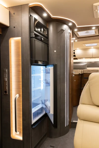 Der Innenraum eines modernen Carthago Chic E-Line-Wohnmobils mit geöffneter Kühlschranktür, die leere Regale zeigt. Warme LED-Beleuchtung hebt die dunklen Holzverkleidungen hervor, und im Vordergrund sind teilweise cremefarbene Ledersitze zu sehen.