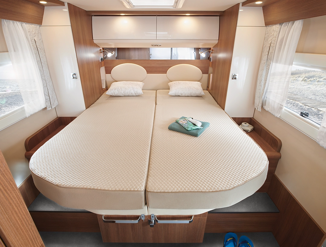 In diesem gemütlichen Schlafzimmer des Carthago C2-Tourer stehen zwei Einzelbetten mit cremefarbener Bettwäsche und Kopfenden nebeneinander. Auf einem Bett liegen gefaltete Handtücher und Seifen, und auf dem grauen Boden neben dem Bett stehen blaue Sandalen. Der Raum ist von Tageslicht durchflutet.