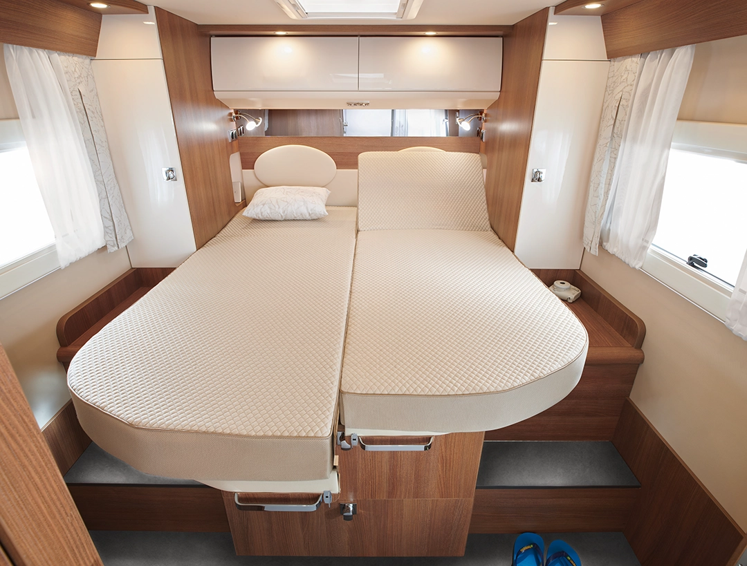 Eine gemütliche Doppelbett-Einrichtung in einem Carthago C2-Tourer-Wohnmobil mit beigen Matratzen, einem Stauraum mit Holzverkleidung darunter, weißen Vorhängen an den Fenstern und blauen Hausschuhen auf dem Boden.