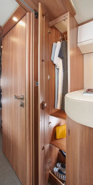 Ein offener Kleiderschrank aus Holz mit einigen Kleidern an Kleiderbügeln, Regale mit einer gelben Tasche und Hüten, daneben ein geschwungenes Bett in einem modernen, kompakten Carthago C2-Tourer-Zimmer.