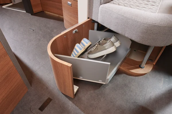 Eine ausziehbare Schuhschublade unter einem gepolsterten Sitz, die geschickt im Stil eines Carthago C2-Tourer gestaltet ist, bietet Platz für zwei Paar Schuhe – ein gestreiftes und ein beiges Paar – in einem modernen Interieur mit Holz- und Grautönen.