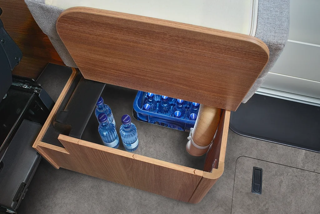 Offenes Staufach aus Holz unter einem Sitz im Carthago C2-Tourer, in dem sich eine Kiste mit Wasserflaschen und ein Stapel Einwegbecher befinden; das Fach befindet sich auf einem mit Teppich ausgelegten Boden neben einem schwarzen Sitzsockel.