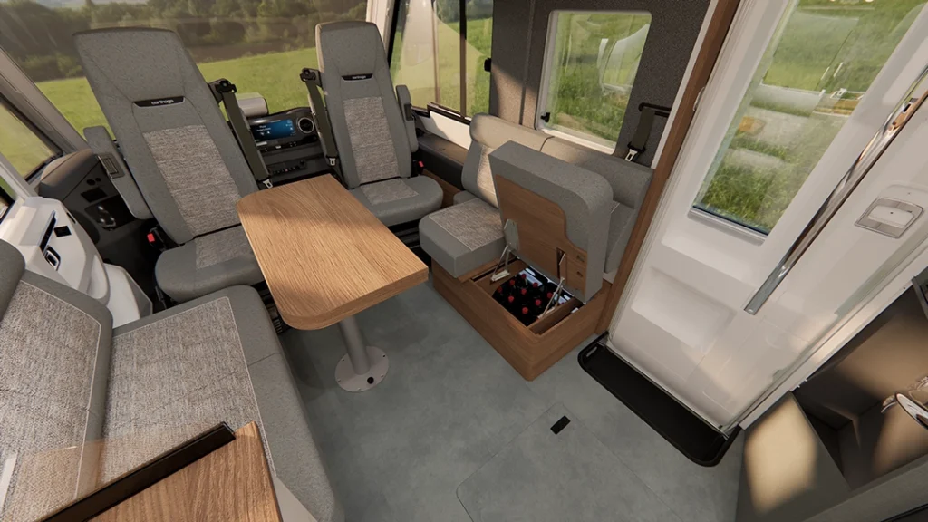 Moderne Wohnmobil-Innenausstattung mit grauen Polstersitzen, einem Holztisch und einem Staufach, das sich öffnen lässt und den Blick auf Getränke freigibt. Große Fenster bieten einen Blick auf die grüne Landschaft draußen. Die Eingangstür befindet sich auf der rechten Seite und unterstreicht das elegante Design des Carthago C2-Tourer.