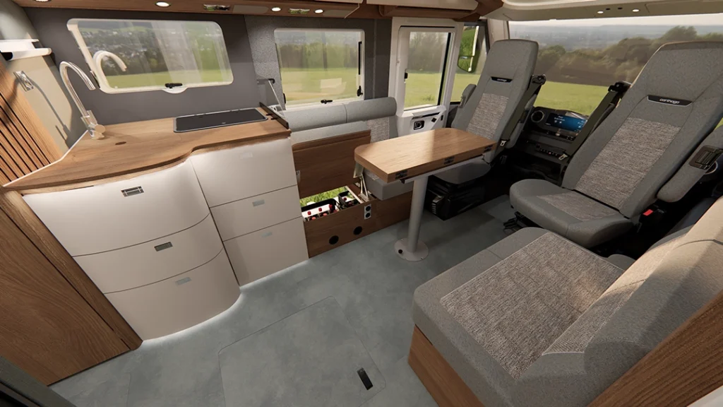 Moderne Innenausstattung eines Wohnmobils mit hellen Holzakzenten, einer kompakten Küche, zwei Drehsitzen, einem kleinen Tisch und großen Fenstern, durch die man die grasbewachsene Landschaft draußen sehen kann. Der Raum ist hell und für die Reise übersichtlich gestaltet, was das durchdachte Design des Carthago C2-Tourer widerspiegelt.
