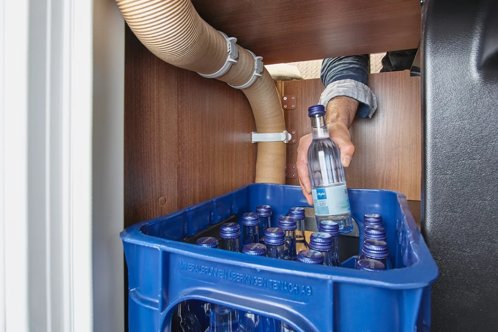 Eine Hand nimmt eine durchsichtige Getränkeflasche aus einer blauen Kiste, die mit ähnlichen Flaschen gefüllt ist und sich unter einem Waschbecken mit freiliegenden Rohren und Schläuchen in der Bordküche eines Carthago C2-Tourer befindet.
