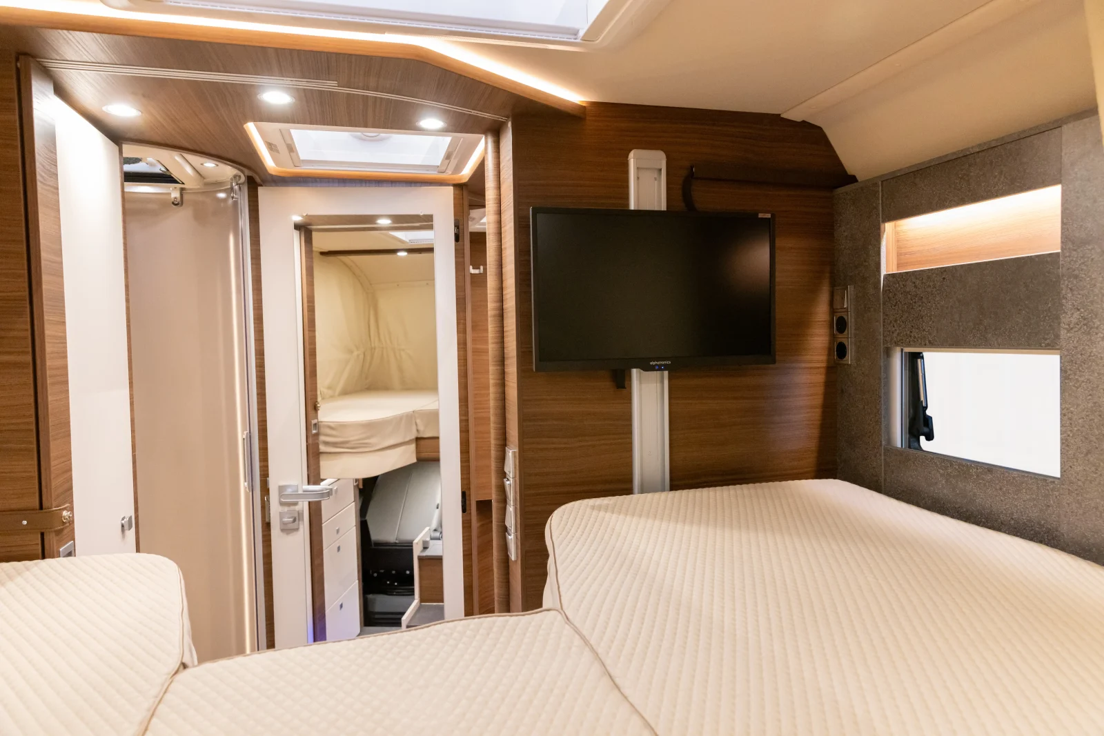 Innenraum eines modernen Carthago Liner for Two-Wohnmobils mit Holzvertäfelung, einem an der Wand montierten Fernseher, zwei Betten mit beiger Bettwäsche und einem kleinen Fenster. Durch die Türöffnung sind ein weiteres Bett und im Hintergrund das Fahrerhaus zu sehen.