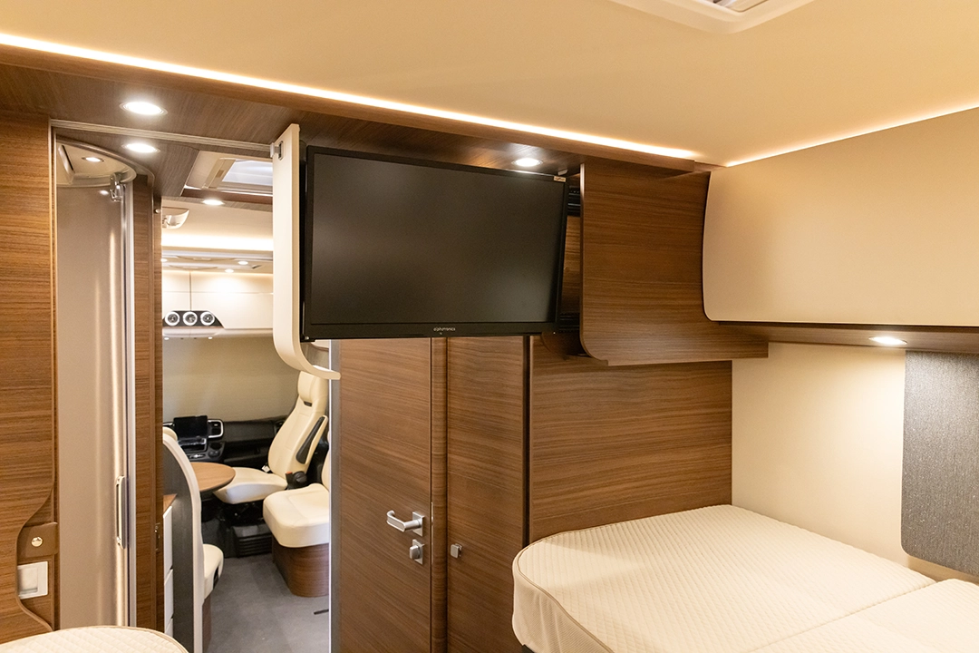 Innenraum eines Carthago Chic E-Line-Wohnmobils mit Holzvertäfelung, zwei Einzelbetten, einem an der Wand montierten Flachbildfernseher und Blick auf den Fahrerbereich und die Drehsitze.