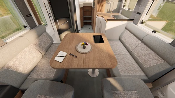 Innenraum eines Carthago C2-Tourer-Wohnmobils mit einem Holztisch, auf dem ein Notizbuch, ein Stift, eine Brille, ein Tablet und eine Blumenvase liegen, umgeben von gepolsterten Sitzbänken, im Hintergrund ist der Küchenbereich zu sehen.