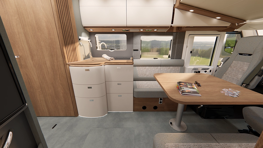 Innenraum eines modernen Carthago C2-Tourer-Wohnmobils mit einer Kochnische, geschwungenen Schränken, einem Esstisch aus Holz mit Zeitschriften und Karten, einer gepolsterten Sitzbank sowie Fenstern mit malerischem Ausblick auf die Landschaft.