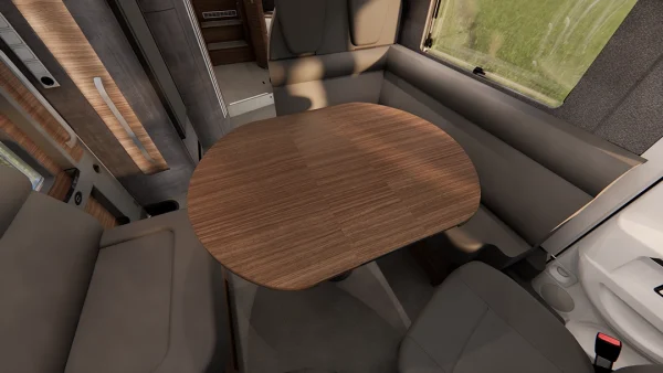 Ein Holztisch, umgeben von grauen Polstersitzen im Inneren eines Carthago Chic E Line-Wohnmobils, mit einem Fenster, durch das man das grüne Gras draußen sehen kann, und im Hintergrund sichtbaren Details der Innenausstattung.