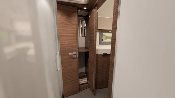 Offener Schrank in einem modernen Wohnmobil oder Wohnwagen der Carthago Chic E-Line mit Holztüren, aufgehängten Hemden, Regalen mit gefalteten Handtüchern und einem kleinen Fenster mit einem Landschaftsbild über dem Regal. Neutrale Farben und kompakte, effiziente Raumnutzung.