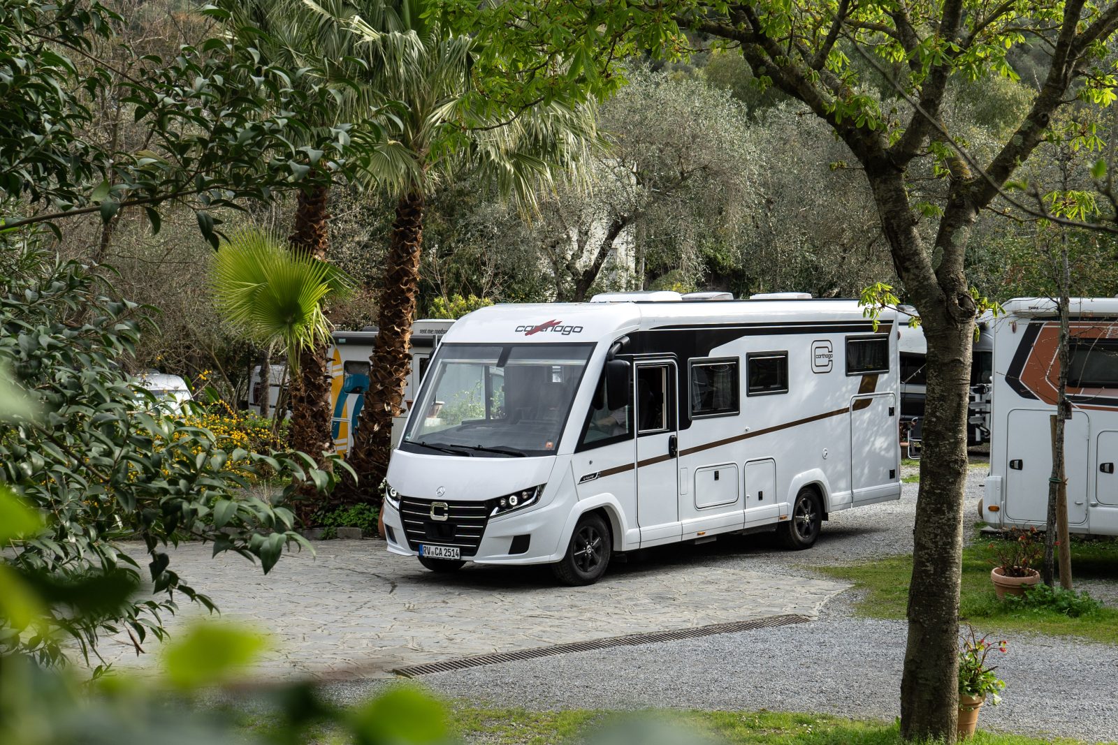 Ein weißer Wohnmobil, der mit den neuesten Modelljahr-Neuheiten ausgestattet ist, steht auf einer gepflasterten Zufahrt, umgeben von grünen Bäumen und Pflanzen in einer Campingplatzkulisse.
