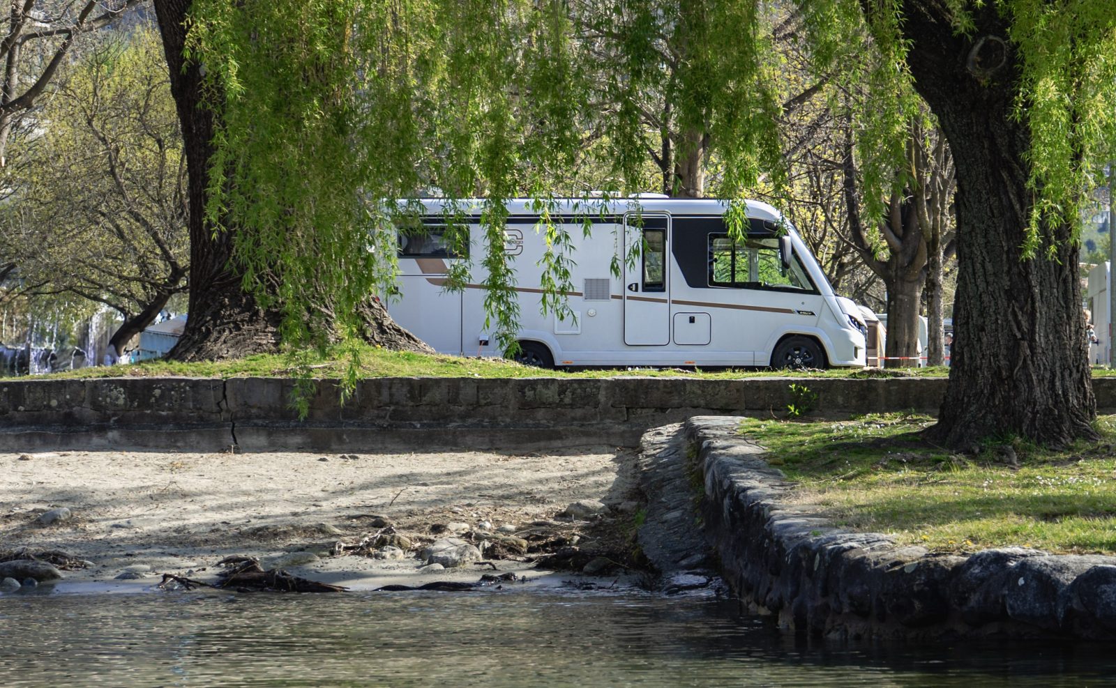 A white motorhome parked beneath deciduous trees near a stone wall, with grass, sand and water in the foreground – the perfect setting to discover all the new features of this model year.