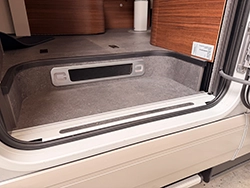 A view of the entrance area of a Carthago chic e-line motorhome, showing a step with a grey non-slip surface, part of the open door and the wood-panelled interior.