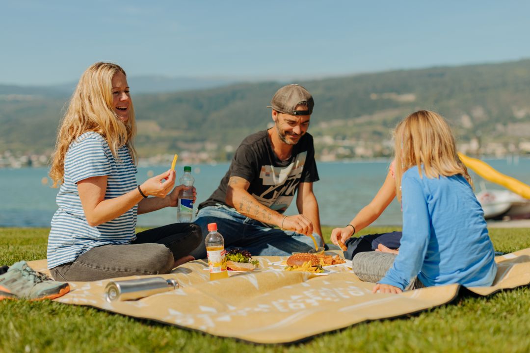 Drei Menschen sitzen auf einer Decke und machen ein Picknick am See. Sie lächeln und genießen gemeinsam das Essen an einem sonnigen Tag, im Hintergrund sind grasbewachsene Hügel und Wasser zu sehen – was an Momente erinnert, die in einem Reisebericht aus Neapel beschrieben werden, in dem sich Freunde versammeln, um inmitten einer wunderschönen Landschaft die einfachen Freuden des Lebens zu genießen.