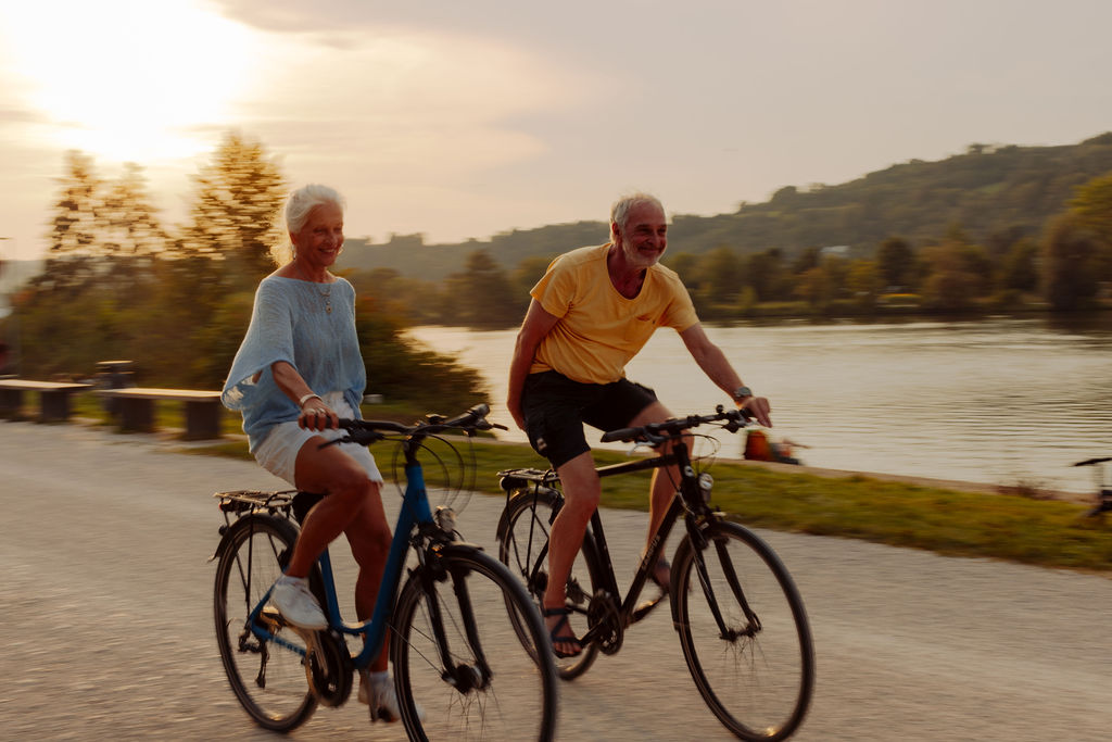 Zwei ältere Menschen fahren bei Sonnenuntergang nebeneinander auf einem Weg in der Nähe eines Flusses Fahrrad, lächeln und genießen die Natur. Im Hintergrund sind Bäume und Hügel zu sehen, die eine einladende Kulisse bilden – perfekt für eine Campingplatzvorstellung.