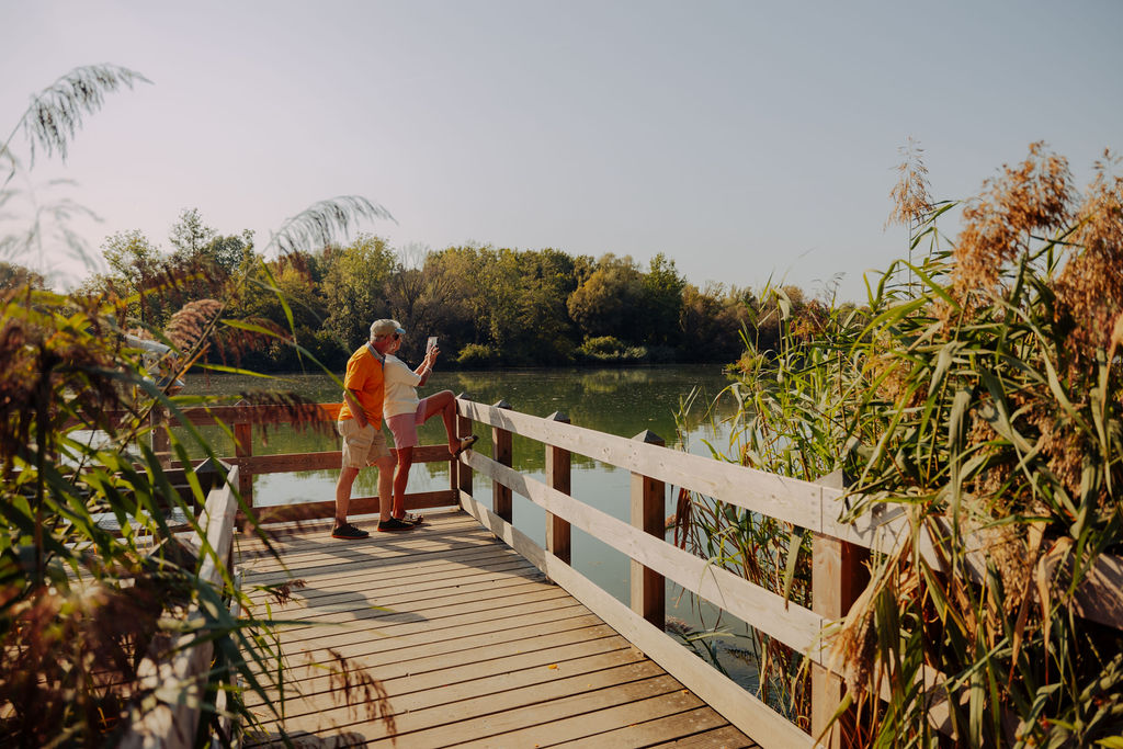 Zwei Menschen stehen auf einem Holzsteg über einem See, umgeben von hohem Gras und Bäumen, und genießen an einem sonnigen Tag die Aussicht – eine perfekte Kulisse für jede Campingplatzvorstellung.
