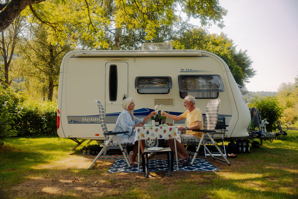 Ein älteres Ehepaar sitzt mit Getränken an einem Tisch vor einem Wohnmobil, lächelt sich an und genießt an einem sonnigen Tag die Natur, umgeben von Bäumen und viel Grün – ein perfekter Moment, der während ihrer Campingplatzvorstellung eingefangen wurde.