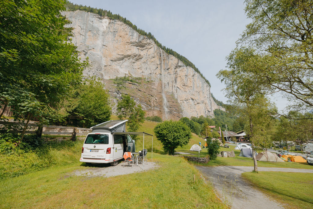 Ein weißer Wohnwagen mit Markise steht auf grünem Rasen neben Campingstühlen und einem Tisch, umgeben von Bäumen und mit einer hohen Klippe im Hintergrund – eine einladende Szenerie, die an einen Reisebericht aus Neapel erinnert, in dem Abenteuer und natürliche Schönheit miteinander verschmelzen.