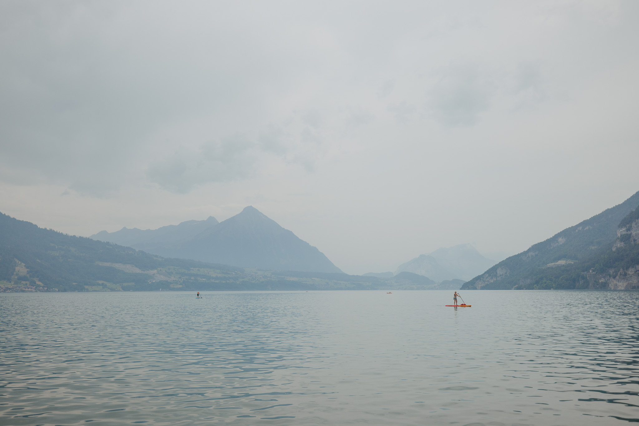 Eine Person paddelt auf einem ruhigen, von Bergen umgebenen See unter bewölktem Himmel und fängt dabei die friedliche Atmosphäre ein, die an Szenen erinnert, wie sie in einem „Reisebericht Neapel“ beschrieben werden. Weiter entfernt sind auf dem Wasser weitere kleine Gestalten zu sehen.