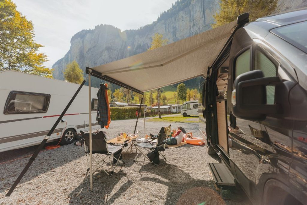 Ein Campingplatz mit einem Wohnmobil und einer Markise, Stühlen, einem Tisch mit Essen und Bergen im Hintergrund. Das Sonnenlicht scheint über die Bäume und die geparkten Wohnwagen und fängt die perfekte Szene für einen Reisebericht über Neapel ein.