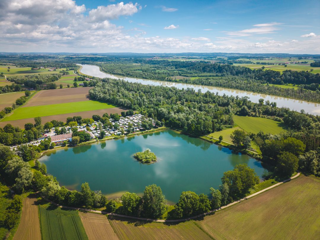 Luftaufnahme eines Sees mit einer kleinen Insel, umgeben von Bäumen, Feldern und einer Reihe von Gebäuden – perfekt für eine Campingplatzvorstellung. In der Nähe fließt ein größerer Fluss unter einem teilweise bewölkten Himmel, am Horizont sind in der Ferne Hügel zu sehen.