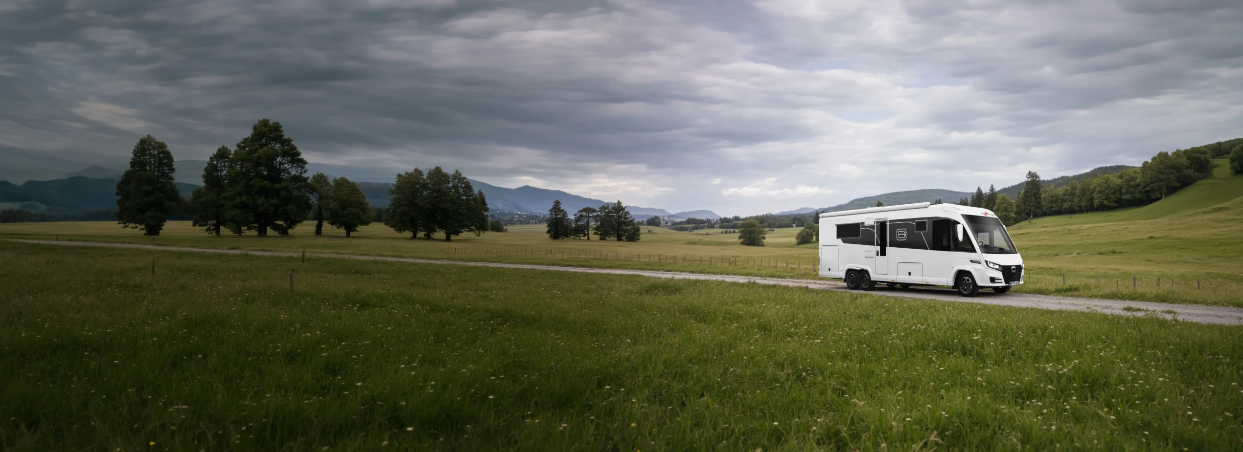 Ein Carthago Liner for Two-Wohnmobil fährt auf einer schmalen Straße durch ein weitläufiges, grünes Feld mit vereinzelten Bäumen unter bewölktem Himmel und vor dem Hintergrund ferner Berge.