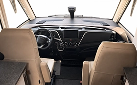A view from the rear of the cab of a beige Carthago chic s-plus motorhome; the steering wheel, the dashboard with a screen and two front seats can be seen. The windscreen is large and clear, and light streams in.
