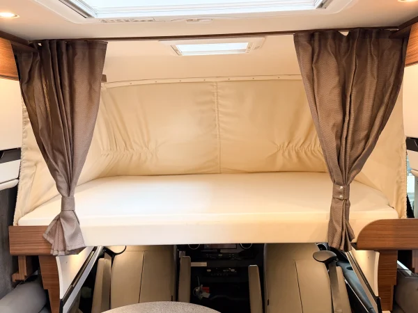 Klappbett mit beiger Matratze und braunen Vorhängen im kompakten Innenraum des Carthago Chic E-Line, das sich über dem Fahrerbereich befindet. Das Bett ist ordentlich bezogen und die Vorhänge sind auf beiden Seiten zurückgebunden.