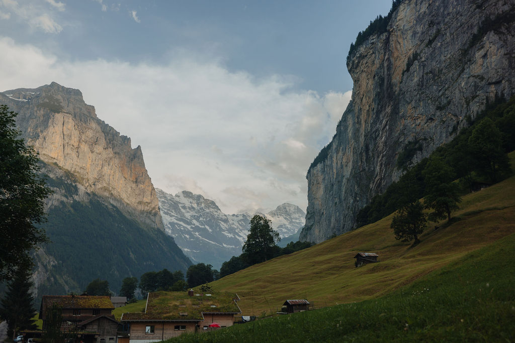 Auf einem grasbewachsenen Hügel am Fuße hoher, steiler Klippen stehen Holzhäuser, im Hintergrund sind schneebedeckte Berge und ein bewölkter Himmel zu sehen – eine Landschaft, die an Szenen aus einem „Reisebericht Neapel“ erinnert, in der natürliche Schönheit auf beschauliche Abgeschiedenheit trifft.