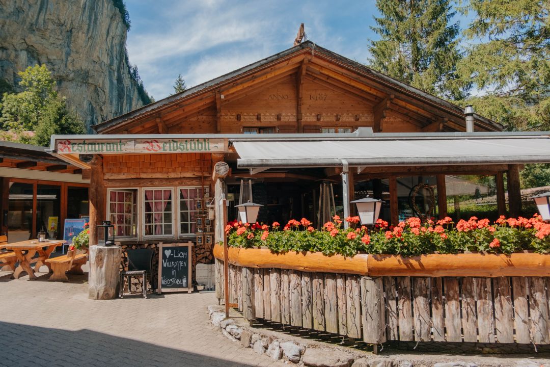 Ein rustikales Restaurant aus Holz mit Blumenkästen voller roter Blüten, Sitzgelegenheiten im Freien und einer Bergkulisse unter blauem Himmel versprüht den Charme, den man oft in einem gut geschriebenen Reisebericht über Neapel findet.