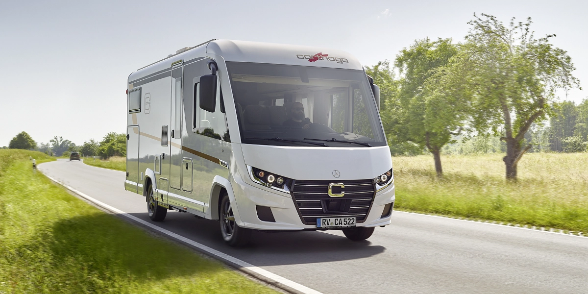 Ein weißes Wohnmobil aus der Serie „Starke Partner“ von Carthago fährt auf einer sonnigen Landstraße, die von grünem Gras und Bäumen gesäumt ist. Das moderne Fahrzeug mit schwarzen Akzenten steht für Qualität und Vertrauen – perfekt unterwegs unter klarem Himmel.