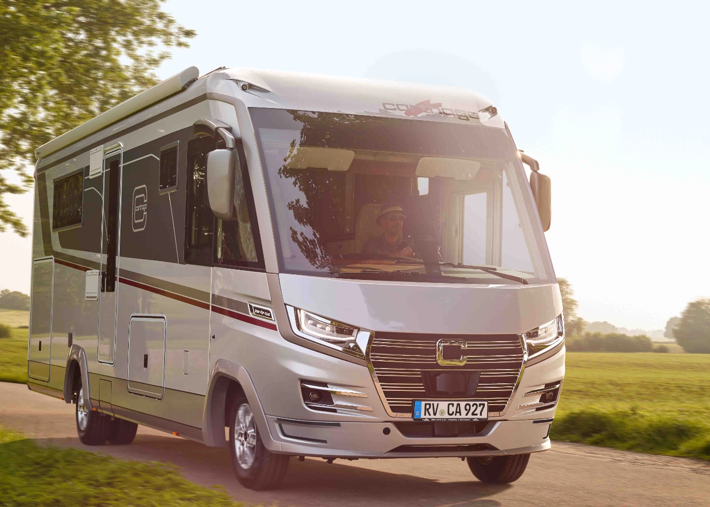 An einem sonnigen Tag fährt ein weißer Wohnmobil auf einer von Gras und Bäumen gesäumten Landstraße entlang, im Hintergrund sind Felder und ein Haus in der Ferne zu sehen – Qualität und Vertrauen, wie sie die starken Partner von Carthago bieten.