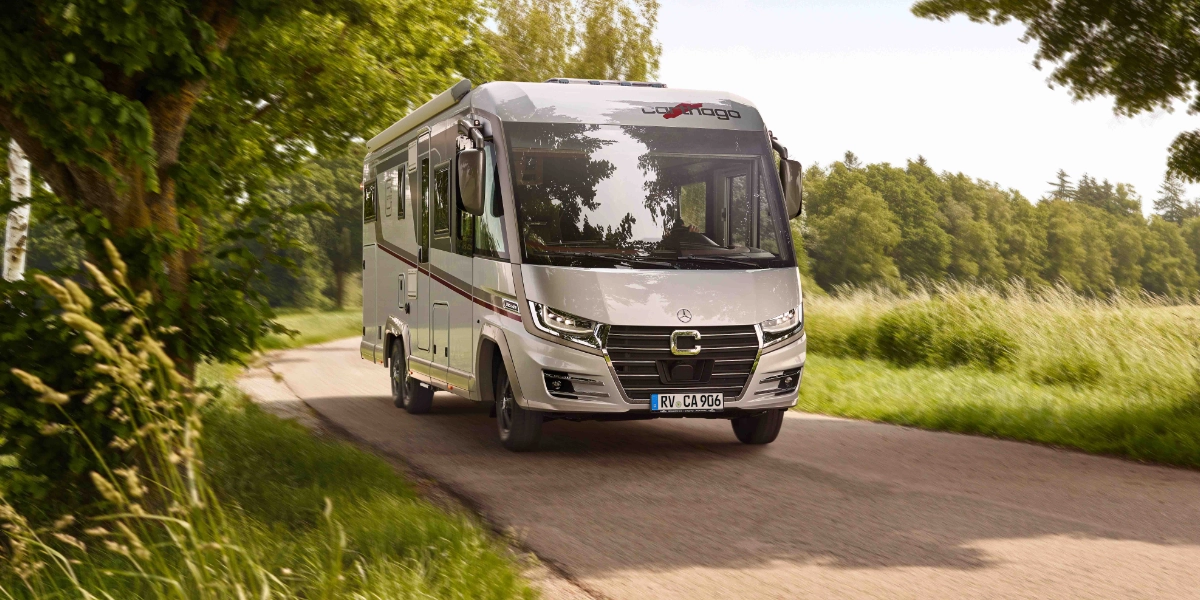 Ein silbernes Wohnmobil fährt unter strahlend blauem Himmel eine schmale Landstraße entlang, umgeben von grünem Gras, Bäumen und Sträuchern – eine perfekte Szene, die die Qualität und das Vertrauen der starken Partner von Carthago widerspiegelt.