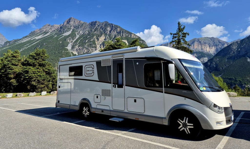 Ein modernes weißes Wohnmobil steht auf einem Parkplatz, im Hintergrund ragen hohe grüne Bäume und schroffe Berge unter einem strahlend blauen Himmel empor – ein Anblick, der das Gefühl von Abenteuer weckt, wie es in jedem Reisebericht über Neapel zu finden ist.