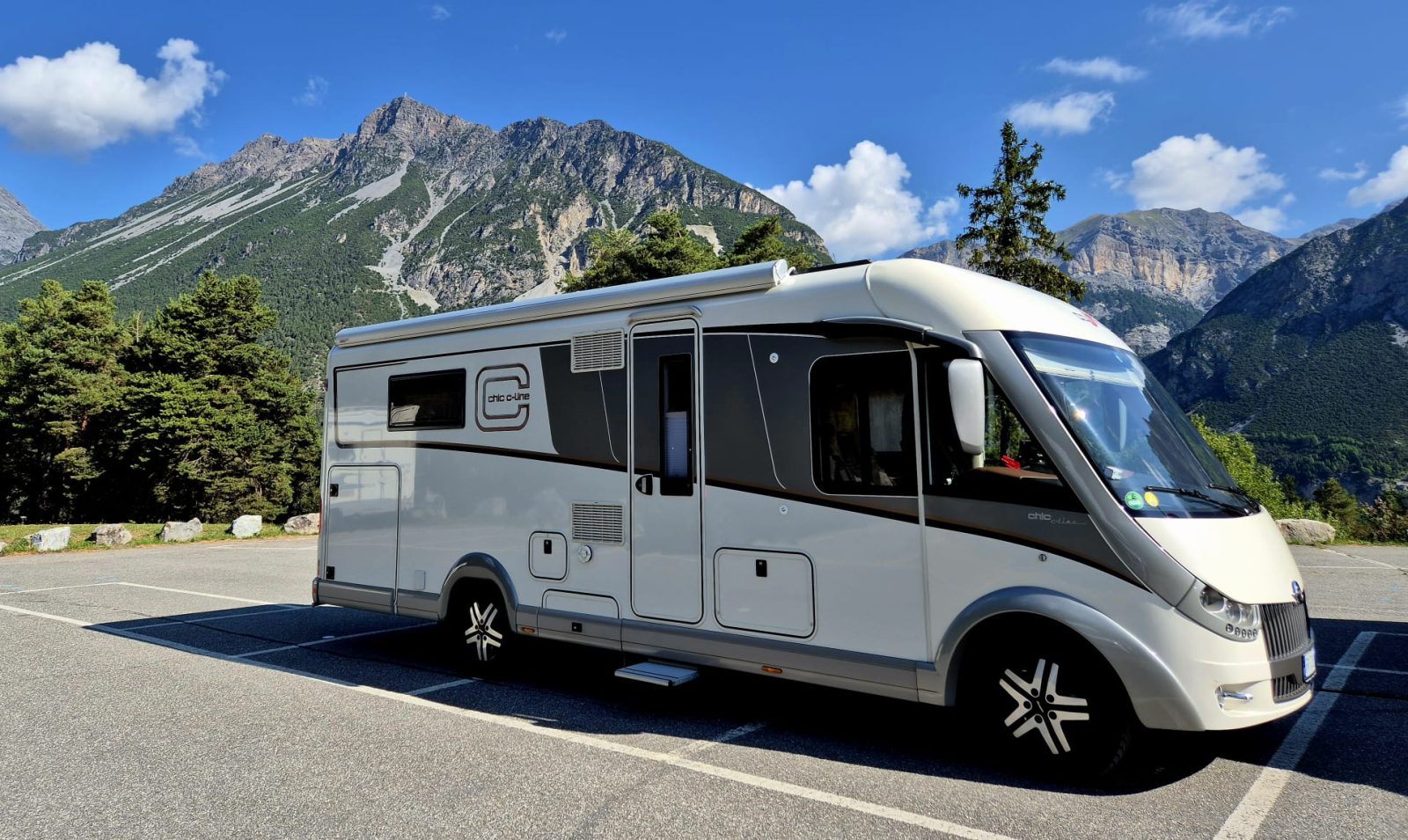 Ein modernes weißes Wohnmobil steht auf einem Parkplatz, im Hintergrund ragen hohe grüne Bäume und schroffe Berge unter einem strahlend blauen Himmel empor – ein Anblick, der das Gefühl von Abenteuer weckt, wie es in jedem Reisebericht über Neapel zu finden ist.