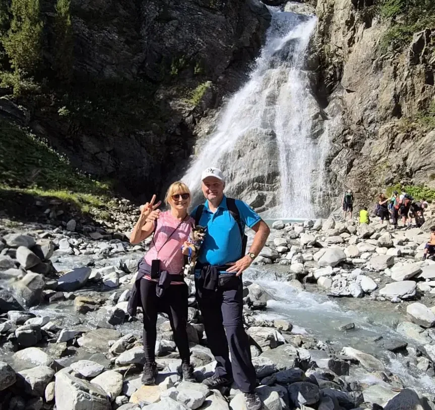 Zwei Menschen stehen auf felsigem Untergrund vor einem hohen Wasserfall, lächeln und posieren für die Kamera. Sie befinden sich im Freien, umgeben von Bäumen, und Sonnenlicht fällt auf die Szene. Im Hintergrund sind weitere Menschen zu sehen, die Momente für ihren eigenen Reisebericht über Neapel festhalten.