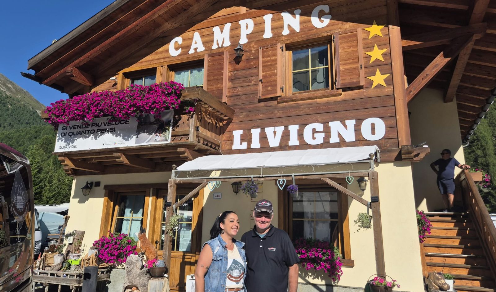 Eine lächelnde Frau und ein Mann stehen gemeinsam vor einem Holzgebäude, an dem Schilder mit der Aufschrift „Camping Livigno“ und zwei gelbe Sterne zu sehen sind. Das Gebäude ist mit Blumenkästen und einem Balkon geschmückt; im Hintergrund sind Berge und blauer Himmel zu sehen – ein Moment, der an Szenen erinnert, wie sie in einem Reisebericht über Neapel beschrieben werden.