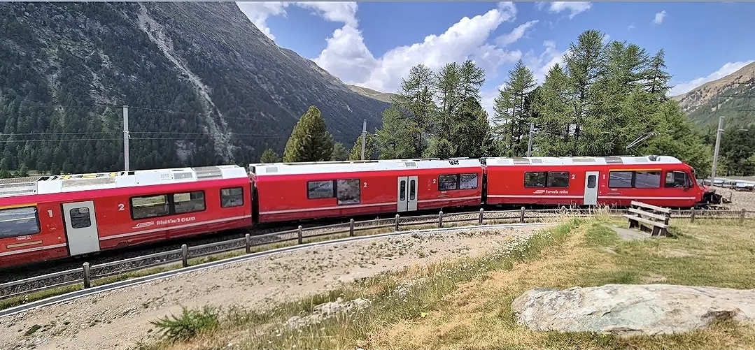 Ein roter Personenzug schlängelt sich auf den Gleisen durch eine bergige Landschaft mit Bäumen und Gras unter einem blauen Himmel mit Wolken und weckt dabei den Entdeckergeist, den man in jedem fesselnden Reisebericht aus Neapel findet.