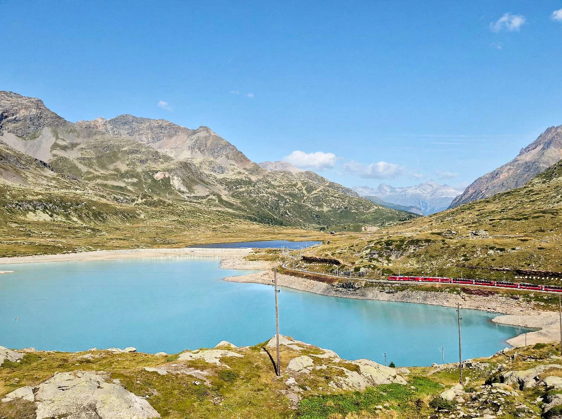 Ein türkisfarbener See, umgeben von Bergen, an dessen Ufer ein roter Zug entlangfährt, unter einem strahlend blauen Himmel – ganz ähnlich wie die Szenen, die in einem „Reisebericht Neapel“ beschrieben werden. Im Vordergrund sind felsiges Gelände und spärliches Gras zu sehen.