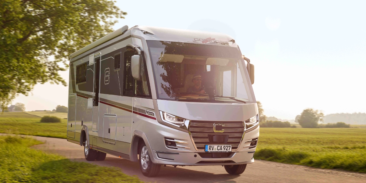 Ein silbernes Reisemobil, das die Werte „Qualität und Vertrauen“ verkörpert, fährt auf einer schmalen Landstraße, die von grünem Gras und Bäumen gesäumt ist, während an einem klaren Tag das Sonnenlicht von rechts hereinströmt – ein echter Beweis für die „Starken Partner von Carthago“.