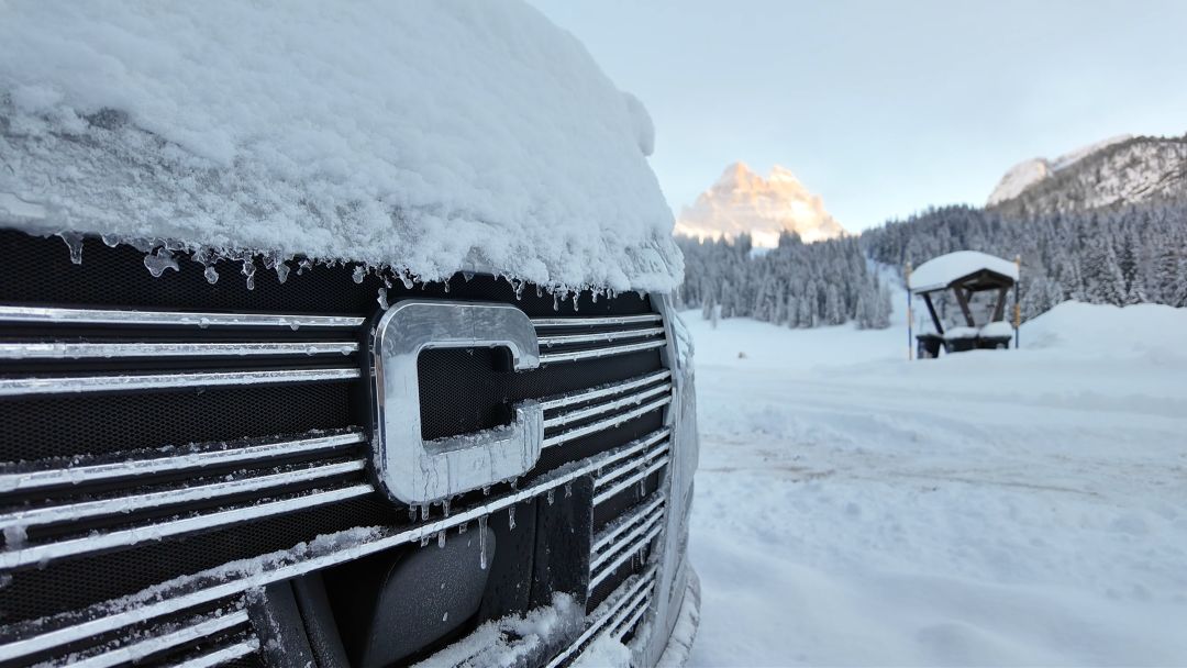 Nahaufnahme eines Kühlergrills mit einem C-Emblem, der mit Schnee und Eis bedeckt ist, vor dem Hintergrund verschneiter Berge auf diesem Wohnmobilstellplatz.