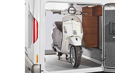 Ein weißer Motorroller ist im Stauraum eines Wohnmobils geparkt, wobei die Tür des Stauraums geöffnet ist, um den Motorroller und einen an der Innenseite der Tür angebrachten Werkzeughalter zu zeigen.