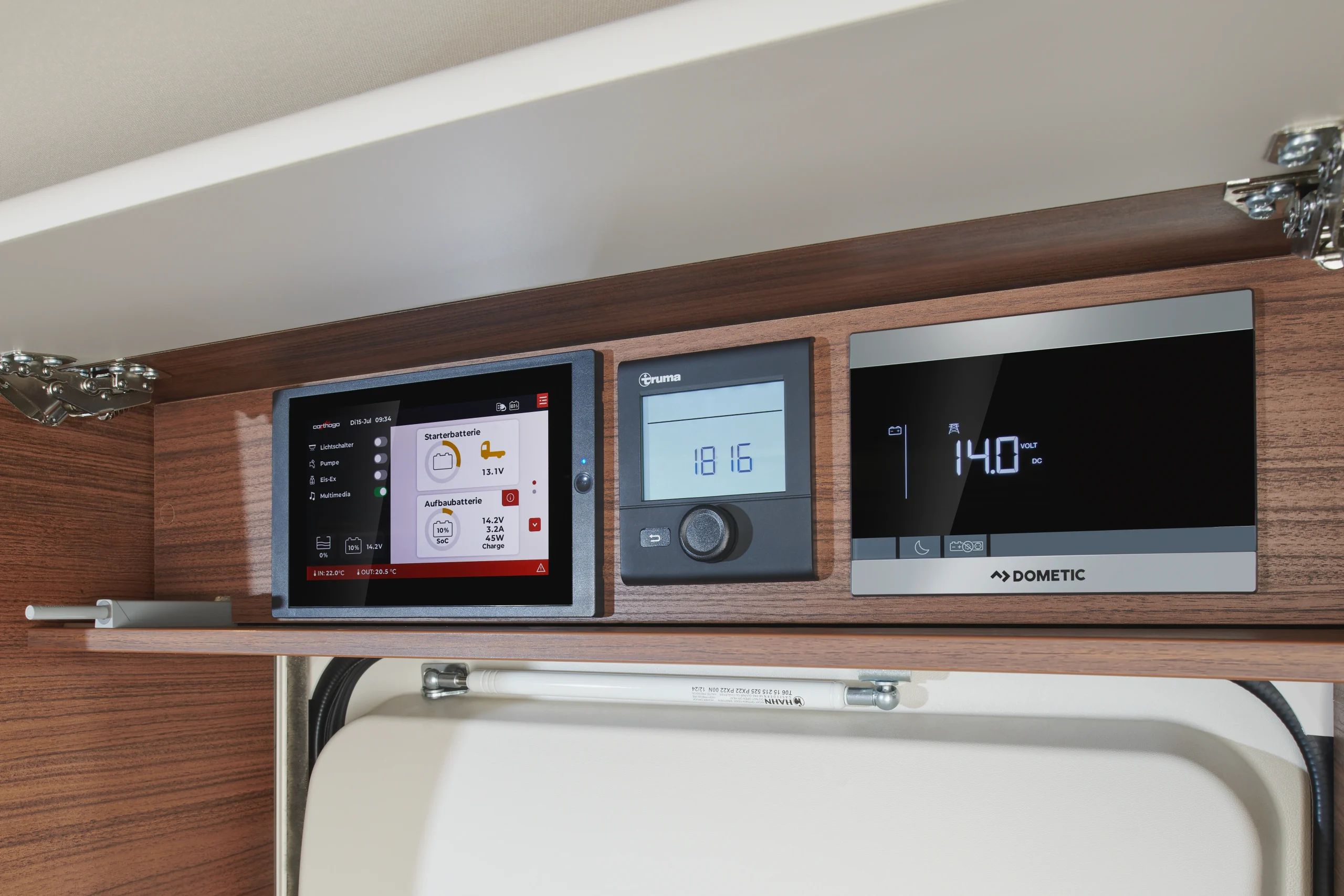 Drei Bedienfelder, eingebaut in einen Holzschrank der Carthago Chic C-Line, darunter eine Touchscreen-Anzeige für den Batteriestatus, ein Truma-Klimaregler und ein Dometic-Batteriemonitor.