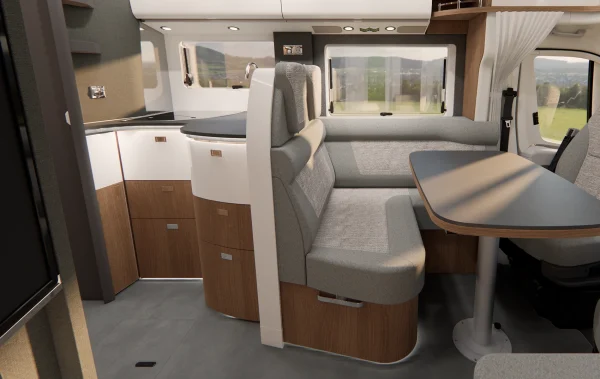 Innenraum eines teilintegrierten Carthago c-tourer-Wohnmobils mit einer grau gepolsterten Sitzbank, einem Esstisch und einer kleinen Küchenzeile mit weißen und holzfarbenen Schränken.