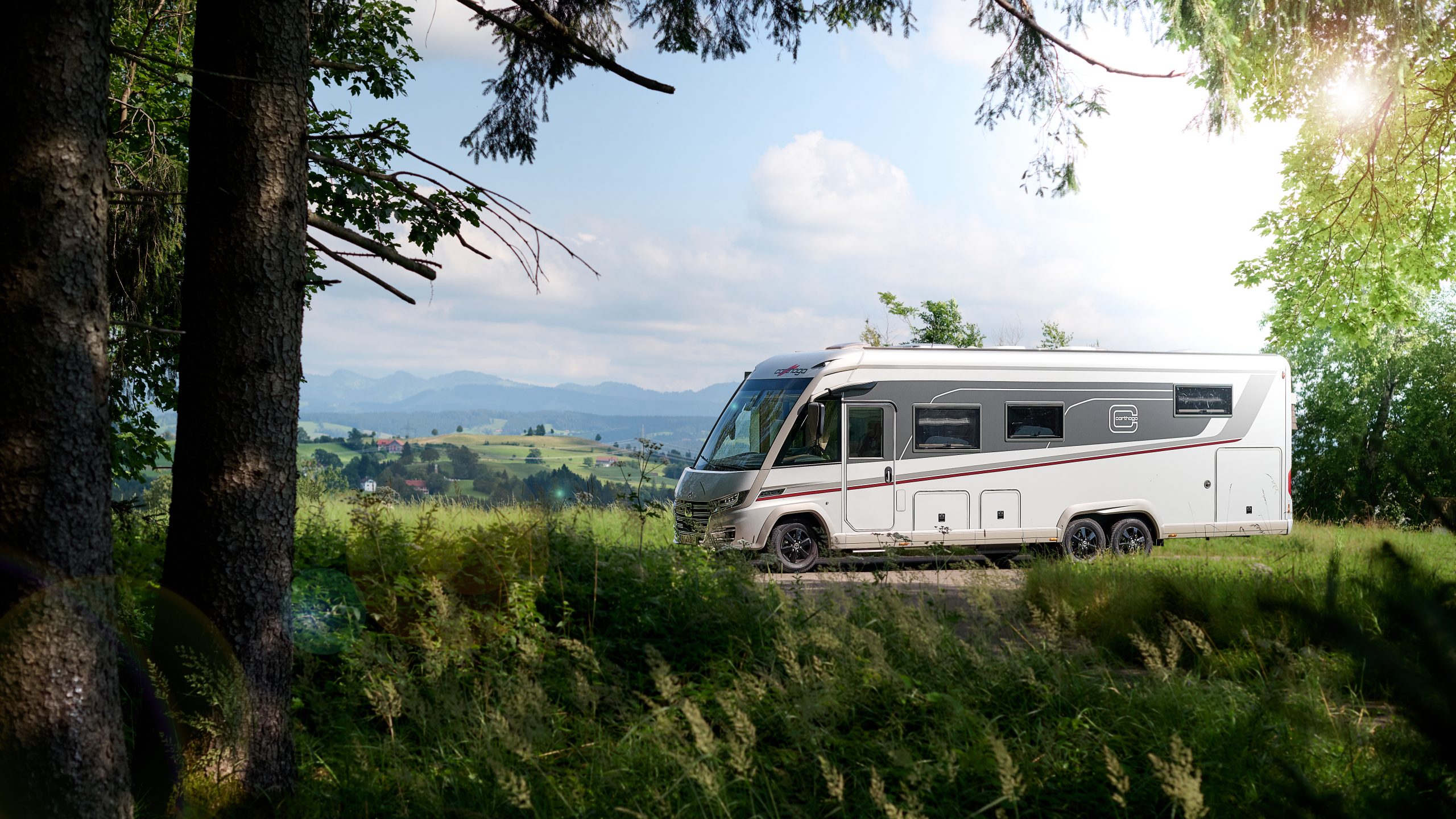 Ein weißes Carthago-Wohnmobil steht auf einem malerischen Wohnmobilstellplatz mit Blick auf sanfte Hügel und Berge in der Ferne.