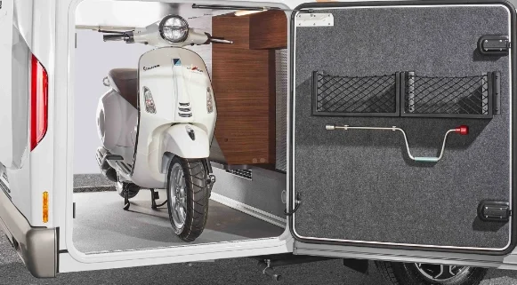 Ein weißer Roller ist im hinteren Staufach eines integrierten Wohnmobils geparkt. Die Tür des Fachs ist geöffnet und gibt den Blick auf den Roller und einige Regale frei.